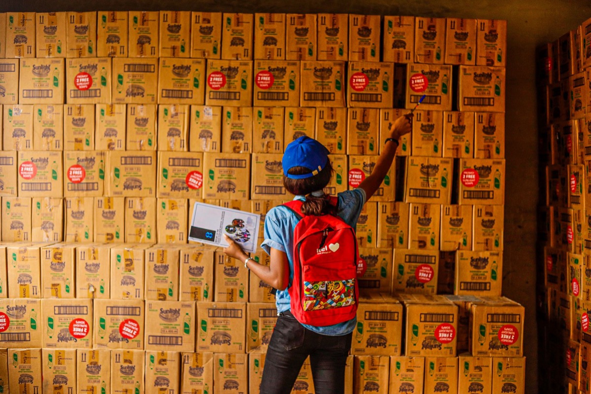 Una persona organizando inventario con un portapapeles en un almacén lleno de cajas.